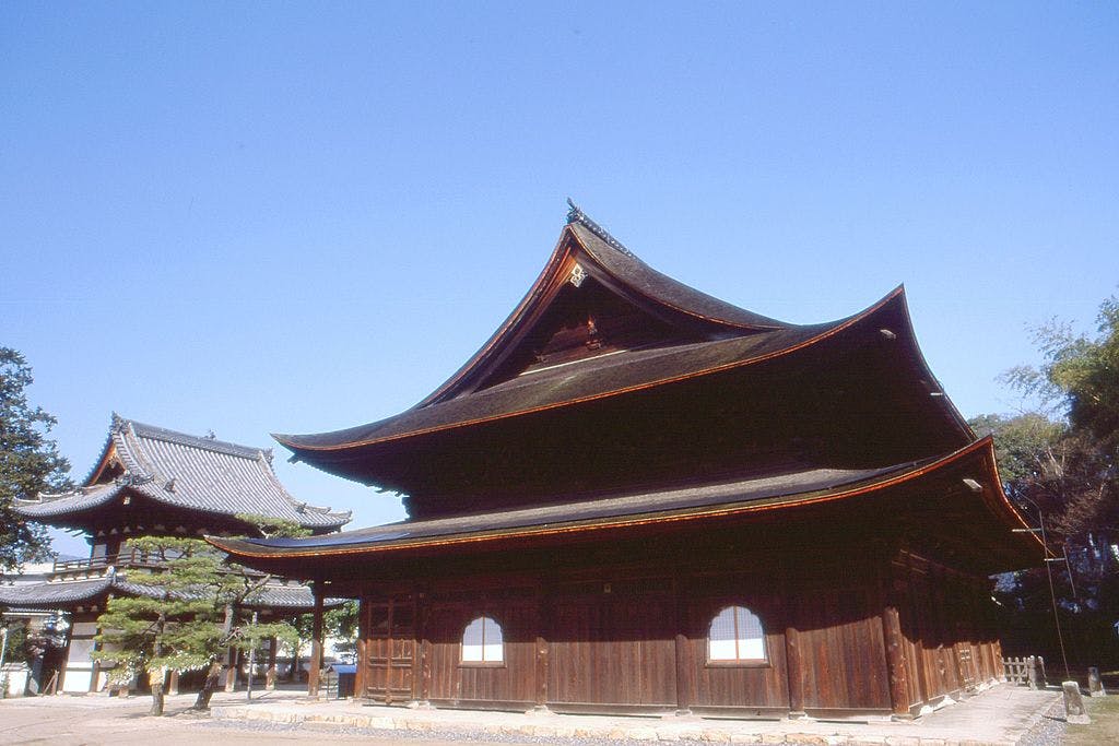 A traditional Japanese wooden temple with a curved, layered roof stands under a clear blue sky, surrounded by trees and another temple building in the background.
