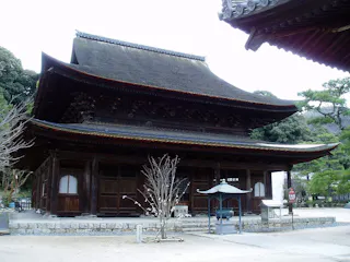 A traditional Japanese wooden temple with a sloped roof sits surrounded by trees and a courtyard, featuring stone lanterns and a leafless tree in front.