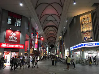 A bustling, covered shopping street in Japan at night, with bright signs for Taito Station arcade and Lawson convenience store. People walk along the wide, well-lit walkway under a high, arched ceiling.