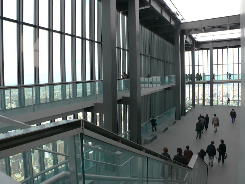 Interior of a tall building with large windows offering a panoramic city view. Multiple levels are visible with people walking and chatting on each floor. The scene includes glass railings, a staircase, and high ceilings, creating an open, modern atmosphere.