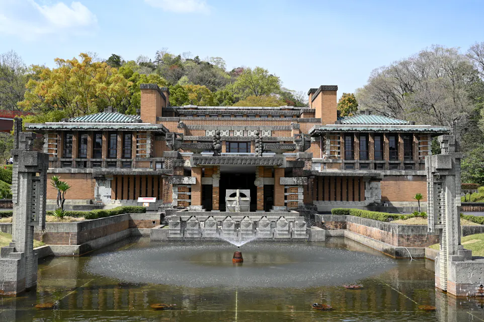 A large brick and stone building with intricate designs, featuring a symmetrically arranged facade. The structure is surrounded by lush greenery and situated behind a serene pond with a central fountain. Trees are visible in the background under a clear blue sky.