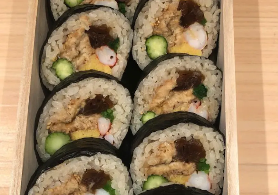A wooden box contains a neatly arranged assortment of sushi rolls. Each roll is wrapped in nori seaweed and filled with rice, egg, shrimp, cucumber, and other ingredients. The sushi rolls are placed closely together, showcasing their vibrant and fresh appearance.