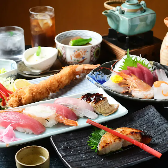 A vibrant Japanese meal spread featuring assorted sushi, sashimi, a bowl of shrimp tempura, a teapot set, and various side dishes. The table also includes a glass of iced tea, a cup of water, and a small bowl of soup, all carefully arranged on a dark surface.