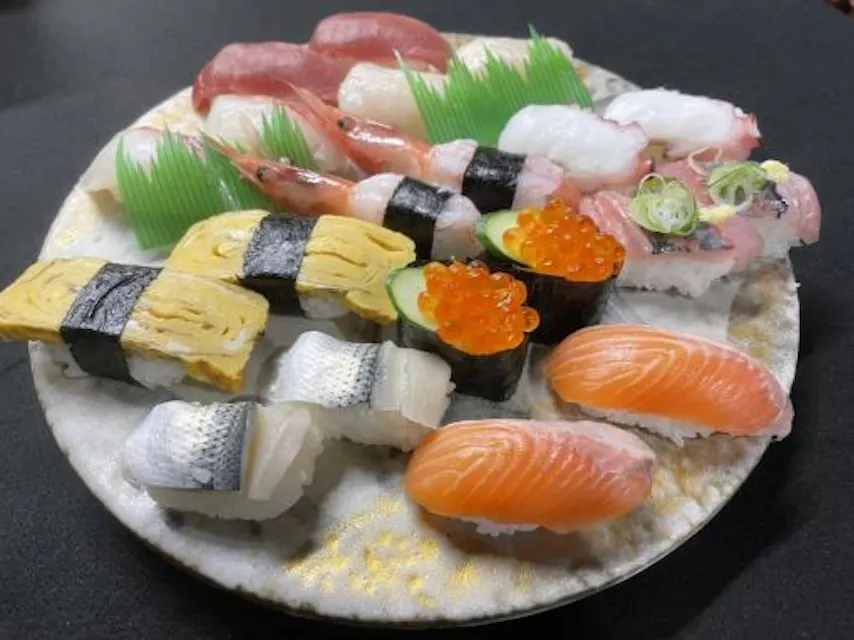 A plate filled with various types of sushi, including nigiri with salmon, squid, shrimp, egg, fish roe, and other assorted seafood. The sushi is arranged neatly on a round plate, garnished with imitation grass separators.
