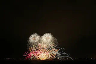Colorful fireworks burst in the night sky above a dark landscape, creating arches and patterns of red, yellow, white, and blue light against a black background.