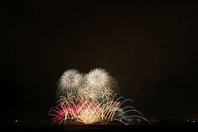 Colorful fireworks burst in the night sky above a dark landscape, creating arches and patterns of red, yellow, white, and blue light against a black background.