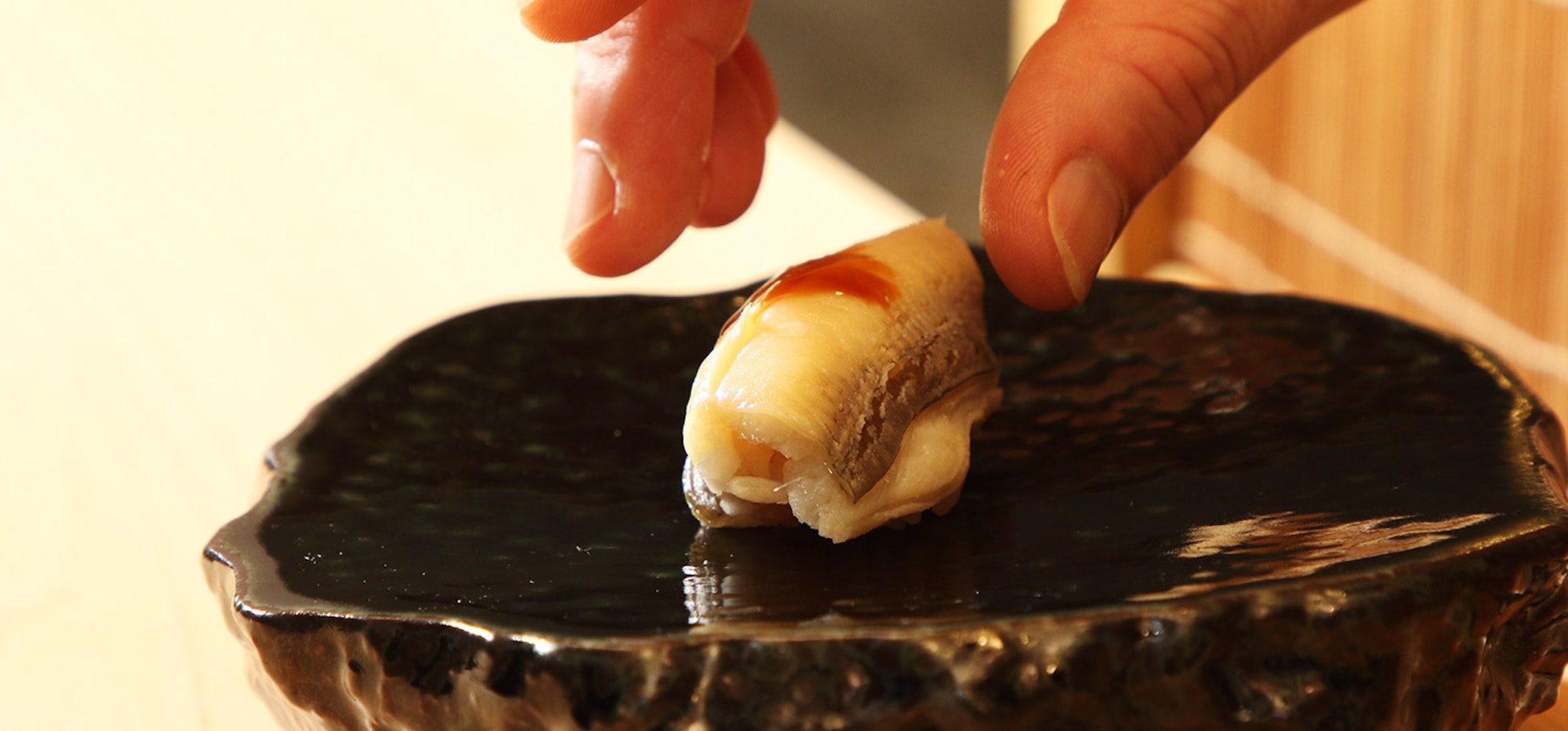 A close-up image of a hand carefully placing a piece of sushi on a dark, glossy plate. The sushi appears to be topped with a slice of fish and a light glaze. The setting has a warm, inviting atmosphere with natural light.