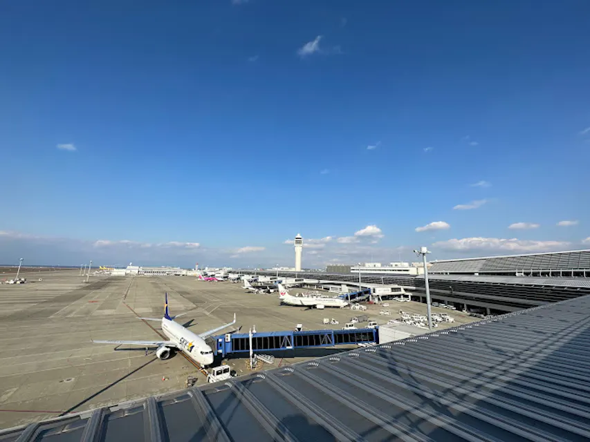 Central Japan International Airport’s Sky Deck A wide-angle view of an airport apron with several airplanes parked at gates. The scene shows a clear, sunny sky with scattered clouds. The control tower is visible in the background, and airport buildings and infrastructure occupy the right side of the image.