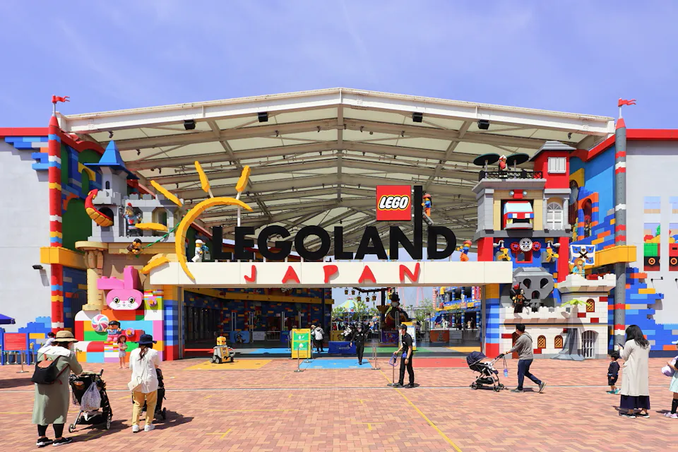 LEGOLAND Japan Entrance to LEGOLAND Japan featuring a colorful, brick-themed façade with various LEGO character sculptures and designs. People are walking and pushing strollers in front of the entrance under a clear sky. The large LEGO logo is prominently displayed above the entrance.
