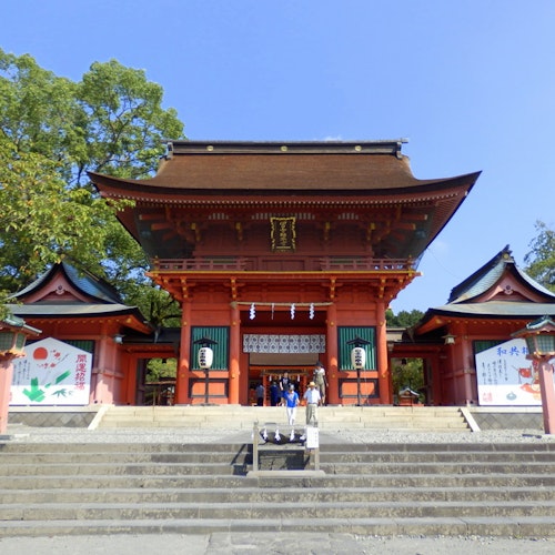 Fujisan Hongu Sengen Taisha Shrine A traditional Japanese shrine with a large red entrance gate, green roofs, and stone steps leading up. Trees and two colorful banners flank the building under a clear blue sky.