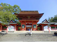 A traditional Japanese shrine with a large red entrance gate, green roofs, and stone steps leading up. Trees and two colorful banners flank the building under a clear blue sky.