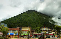 A lush green mountain rises behind traditional Japanese buildings and shops, with low clouds and mist partially covering the peak. People and a white van are visible on the street in the foreground.