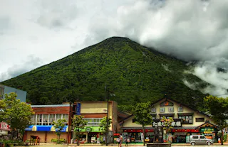 A lush green mountain rises behind traditional Japanese buildings and shops, with low clouds and mist partially covering the peak. People and a white van are visible on the street in the foreground.