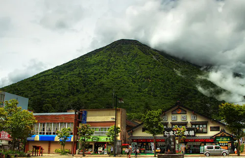 A lush green mountain rises behind traditional Japanese buildings and shops, with low clouds and mist partially covering the peak. People and a white van are visible on the street in the foreground.