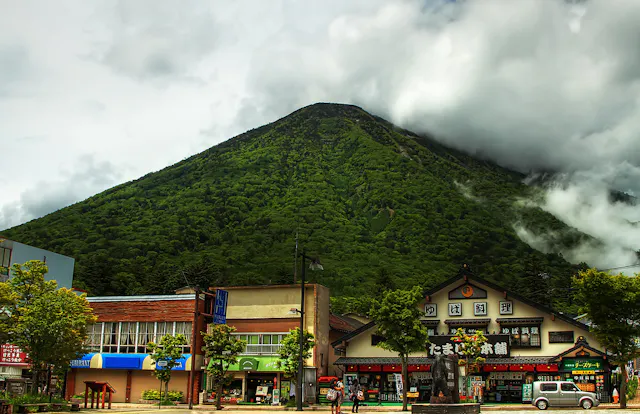 A lush green mountain rises behind traditional Japanese buildings and shops, with low clouds and mist partially covering the peak. People and a white van are visible on the street in the foreground.