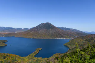 A large, calm blue lake is surrounded by green and autumn-colored trees, with a tall, cone-shaped mountain rising in the background beneath a clear blue sky.