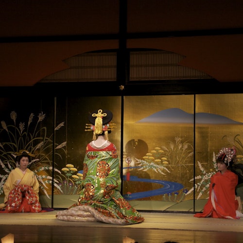 Edo Wonderland Nikko Edomura Three people in traditional Japanese attire are on stage. The center person wears an elaborate green and gold kimono and headpiece, flanked by two others in red robes, kneeling before a painted gold folding screen.