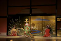 Three people in traditional Japanese attire are on stage. The center person wears an elaborate green and gold kimono and headpiece, flanked by two others in red robes, kneeling before a painted gold folding screen.