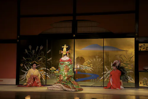 Three people in traditional Japanese attire are on stage. The center person wears an elaborate green and gold kimono and headpiece, flanked by two others in red robes, kneeling before a painted gold folding screen.
