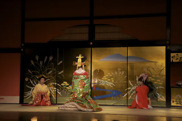 Three people in traditional Japanese attire are on stage. The center person wears an elaborate green and gold kimono and headpiece, flanked by two others in red robes, kneeling before a painted gold folding screen.