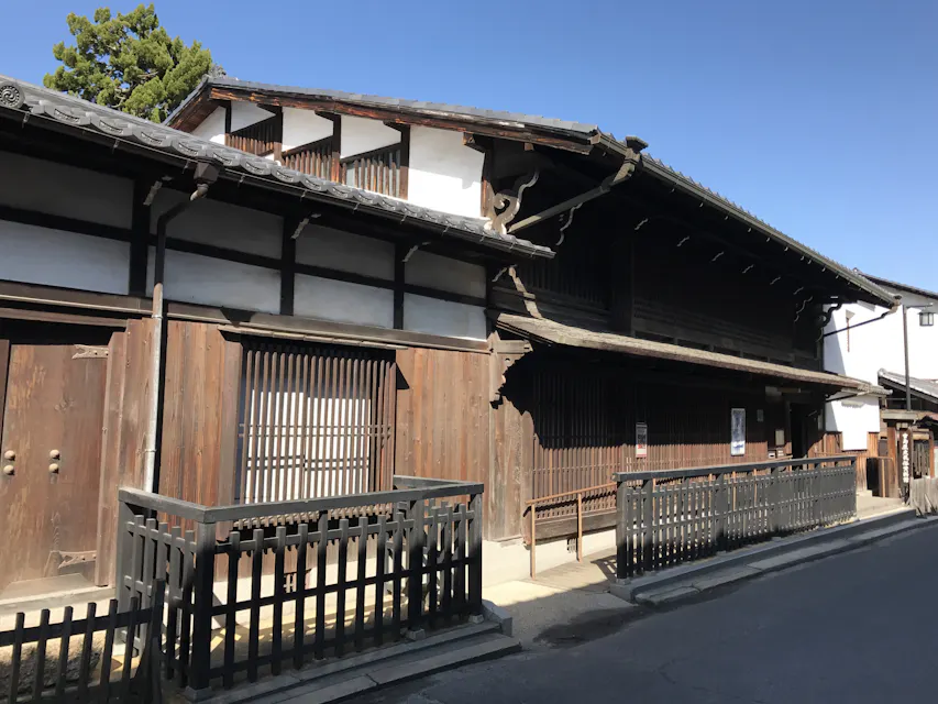 A traditional wooden Japanese building with a tiled roof and wooden lattice windows stands along a quiet street under a clear blue sky. The structure features wooden fences and is shaded by the afternoon sun.