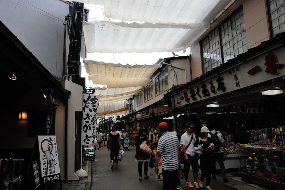 A bustling market street with people walking under a canopy of white tarps. Shops on both sides display various goods, and signs with Japanese characters hang prominently. The atmosphere is lively and shaded from the light.