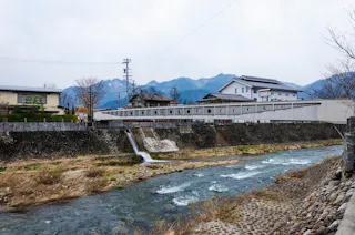 A shallow river flows over rocks with a concrete embankment on both sides, residential houses and a modern building in the background, and distant mountains under a cloudy sky.