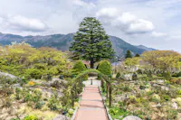 A brick pathway with a green archway leads through a garden filled with bushes and trees, with mountains and a partly cloudy sky in the background. A tall tree stands prominently at the path's end.