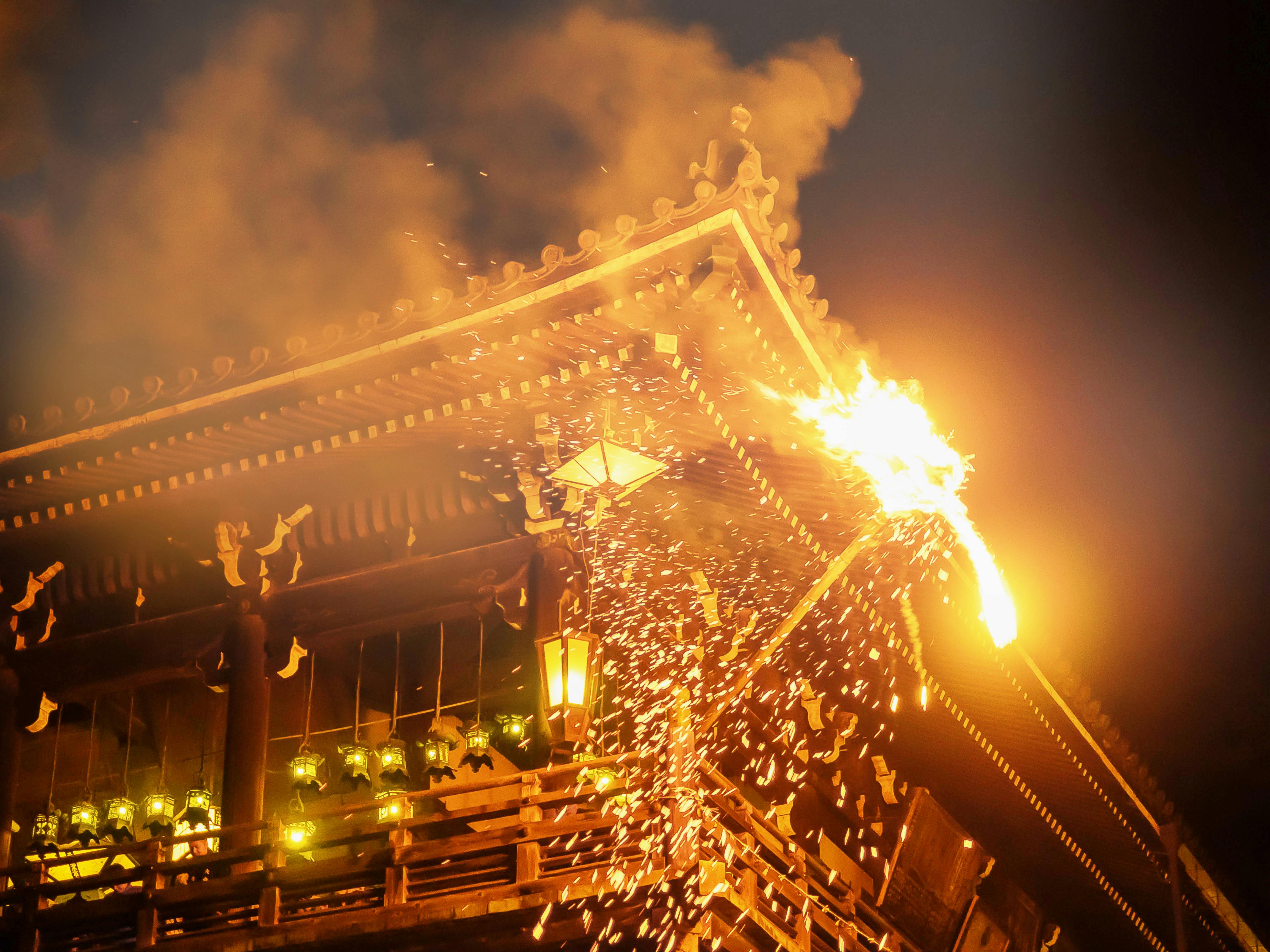 A traditional Japanese wooden temple is shown at night with a large fiery torch, sending sparks and smoke into the air during an annual festival or ritual. Lanterns illuminate the lower level of the building.