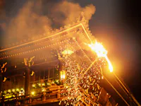 A traditional Japanese wooden temple is shown at night with a large fiery torch, sending sparks and smoke into the air during an annual festival or ritual. Lanterns illuminate the lower level of the building.