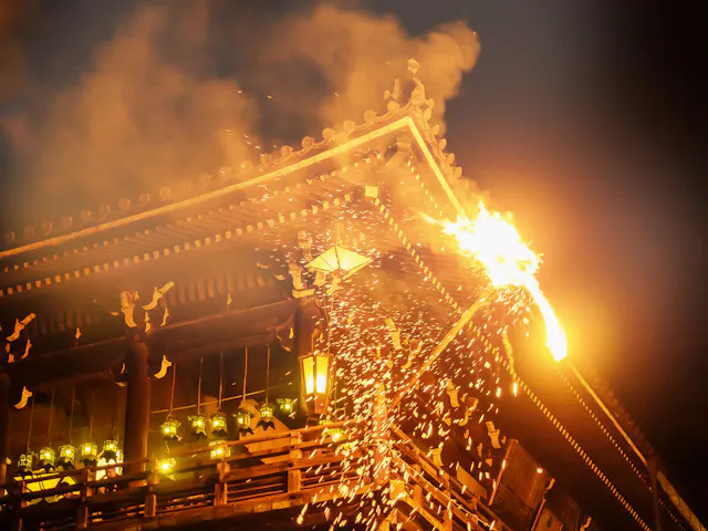 A traditional Japanese wooden temple is shown at night with a large fiery torch, sending sparks and smoke into the air during an annual festival or ritual. Lanterns illuminate the lower level of the building.