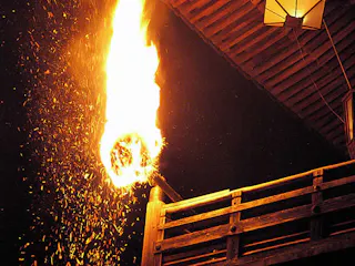 A large, flaming torch emits bright sparks near a wooden balcony at night, with part of the roof and a lantern visible above. The scene is illuminated by the intense fire against the dark background.