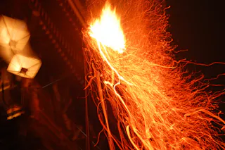 Bright orange sparks and trails of fire shoot through the night sky, creating dynamic, swirling patterns against a dark background, with some blurred lights visible to the left.