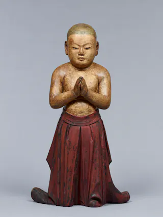 A painted wooden statue of a kneeling child with hands pressed together in prayer, wearing a red skirt-like garment, with a calm expression and a shaved head, set against a plain gray background.