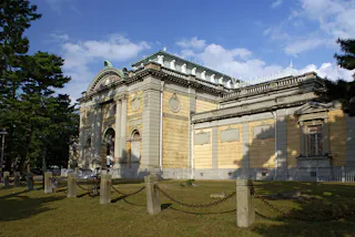 Exterior view of a grand, historic stone building with ornate architectural details, arched windows, and a decorative roof, surrounded by grass, trees, and a chain barrier on a sunny day.