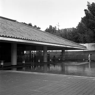 A black-and-white photo of a modern building with a sloped tile roof, large pillars, and glass walls, situated beside a reflecting pool; trees and hills are visible in the background.