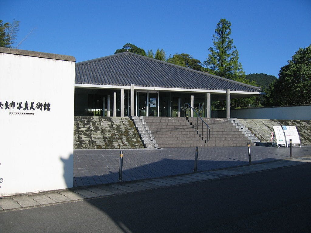 A modern building with a sloped roof, wide steps, and glass entrance stands behind a white wall with Japanese writing. Trees and greenery surround the structure under a clear blue sky.