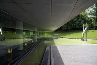 A modern building with large glass windows reflects a green lawn and sculptures. The structure has a cantilevered roof above a shallow reflecting pool, and trees are visible in the background.