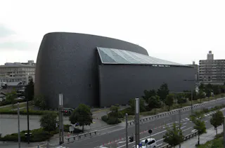 A modern, oval-shaped building with dark, shingled exterior walls and a large slanted glass roof sits beside a road lined with trees and urban buildings in the background.