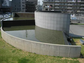Curved, tiered stone water feature with shallow pools and cascading water, set in an urban park area; a person sits near the base, and apartment buildings and parked cars are visible in the background.