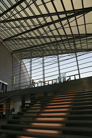 A modern interior with a wide staircase and large glass windows, sunlight casting shadows on the steps, and a person standing on the upper level near the railing.