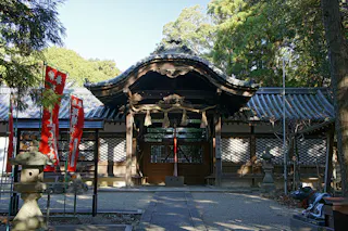 A traditional Japanese shrine entrance with an ornate wooden roof, flanked by red banners and stone lanterns, surrounded by trees and a wooden fence under clear daylight.
