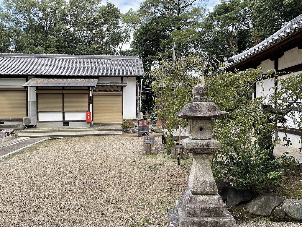 A traditional Japanese courtyard with a gravel ground, stone lantern, trees, and two buildings with tiled roofs and closed shutters. A small red fire extinguisher box is mounted on one building.