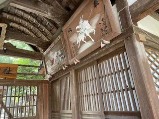 Wooden panels with traditional Japanese artwork and calligraphy hang above a wooden lattice wall in a shrine. One panel features a samurai on horseback, and the other shows a group scene. Lush greenery is visible outside.