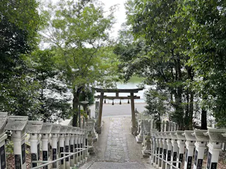 A stone path lined with lanterns leads down steps through lush green trees to a traditional Japanese torii gate, with water and more greenery visible in the background.