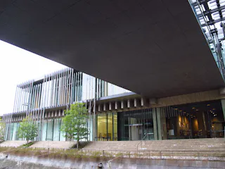 A modern building with large glass windows, vertical metal accents, and an overhanging roof. There are steps, small trees, and a water feature in the foreground. The interior is brightly lit and visible.