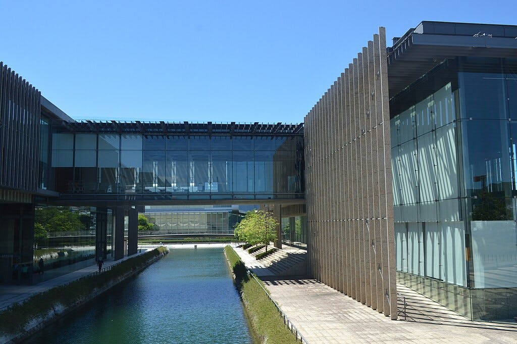 A modern glass and steel building spans over a narrow canal, with clear blue skies above and reflections visible on the water and glass walls. Sunlight casts shadows on the paved walkway beside the canal.