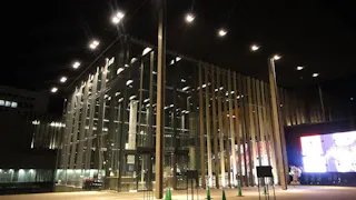 A modern glass building is illuminated at night, with vertical wooden beams, bright lights, and a large screen displaying images outside. Green traffic cones line the walkway at the entrance.