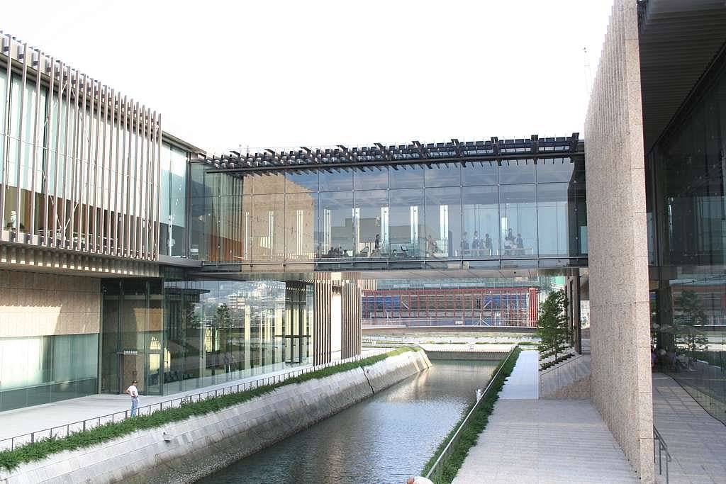 A modern building with glass walls and a sky bridge spans over a narrow canal, with sunlight reflecting off the water and architectural details visible throughout the structure.