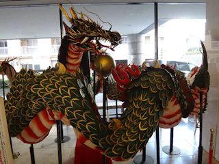 A large, ornate dragon sculpture with green scales, gold accents, and red details is displayed indoors by a window, with its mouth open and a silver sphere in front of it.